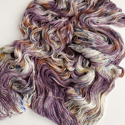 Yarn has been unfurled from its skein and lies across a table. It has been hand dyed using purple, brown and blue dyes. 