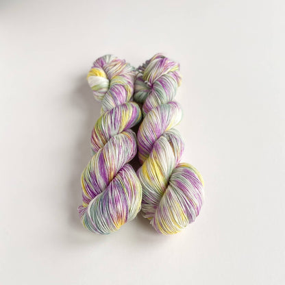 Two skeins of hand dyed yarn on a white table top. The yarn has been dyed with purple, green and yellow dyes. 