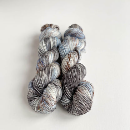 Two skeins of hand dyed yarn on a white table top. The yarn has been dyed with blue, brown and grey dyes. 