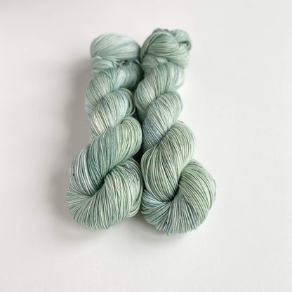 Two skeins of hand dyed yarn on a white table top. The yarn has been dyed with green dyes. 