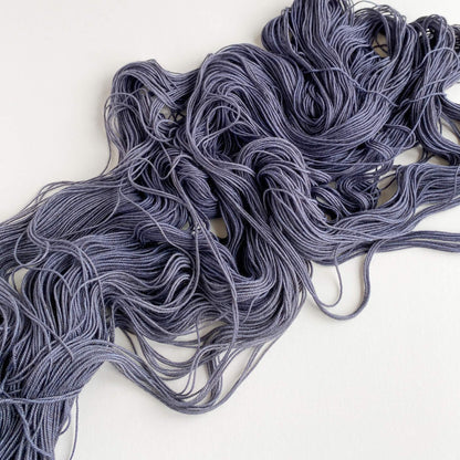 A grey skein of hand dyed yarn lies unfurled on a white table top. 