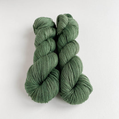 Two skeins of hand dyed yarn on a white table top. The yarn has been dyed with green dyes on a yak base.