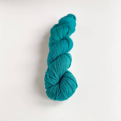 A skein of hand dyed yarn on a white table top. The yarn has been dyed with a teal green dye.