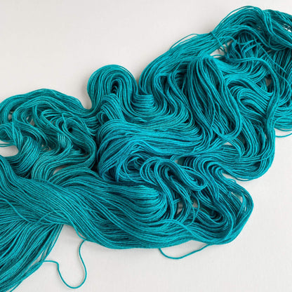 A teal green skein of hand dyed yarn, unfurled, lies on a white table top.