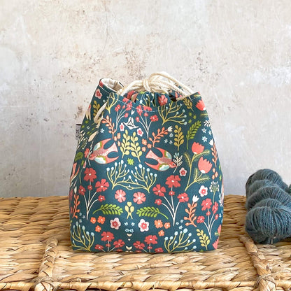 A project bag sits on a wicker chest. It is designed to hold knitting and crochet projects and is made using a dark green nature themed fabric. 