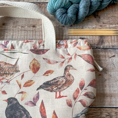 This image shows a close-up of a large knitting project bag with a woodland bird and leaf design in soft, earthy tones. The bag features sturdy fabric handles and a drawstring closure. Nearby, there are skeins of yarn and knitting needles on a wooden surface.
