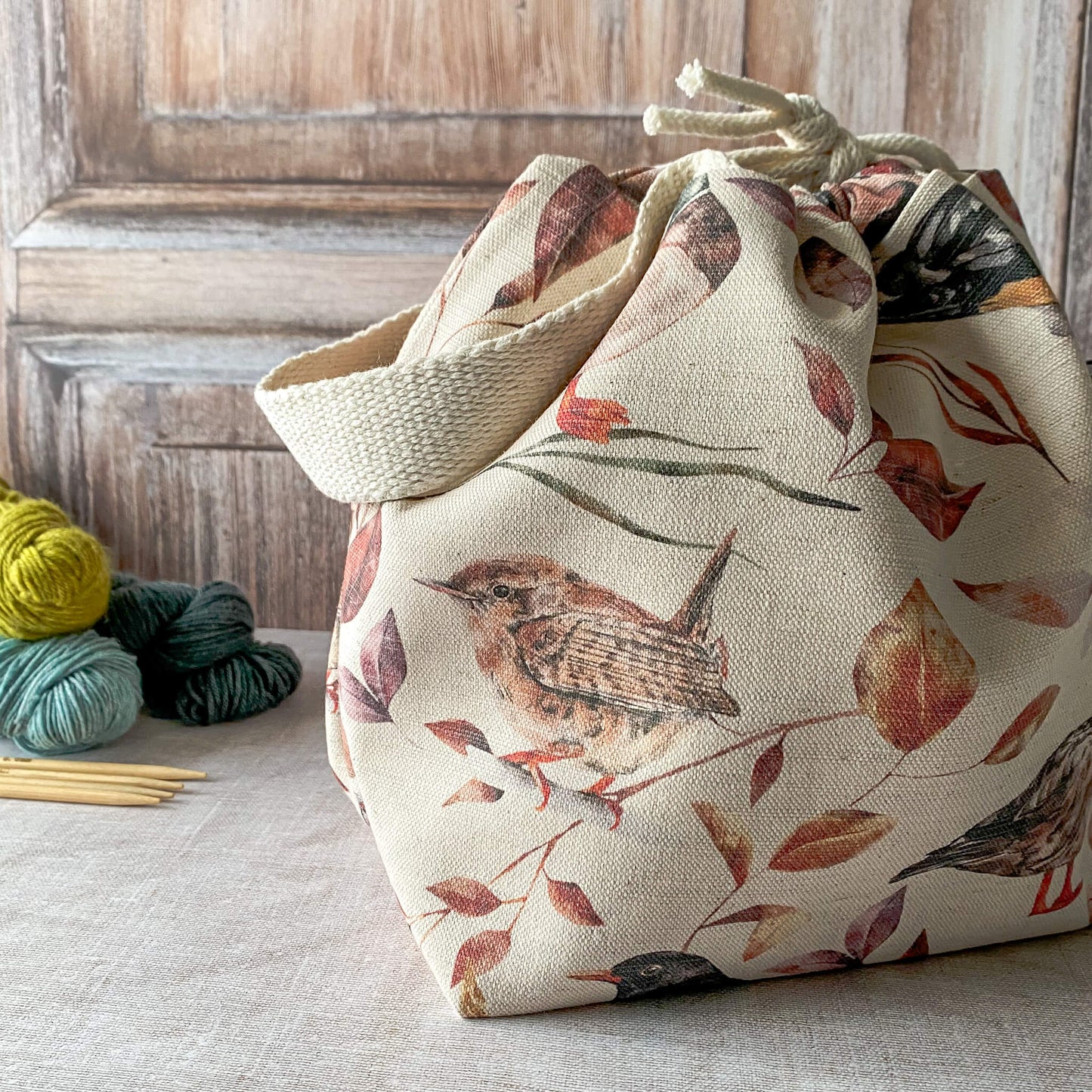 A medium-sized knitting project bag with a delicate woodland bird design in soft, earthy tones. The bag features a drawstring closure with a sturdy handle. Next to it, you can see skeins of yarn and knitting needles placed on a wooden surface.