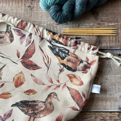 A close-up view of a medium-sized knitting project bag adorned with various birds and autumn leaves. The bag has a drawstring closure and a fabric handle. Visible nearby are knitting needles and yarn in calming tones, set on a wooden background.