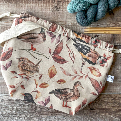 A medium-sized bag with a gentle woodland print featuring birds and colorful leaves. The bag is cinched at the top with a drawstring, and a sturdy handle is visible. It’s placed on a wooden surface with knitting needles and skeins of yarn beside it.