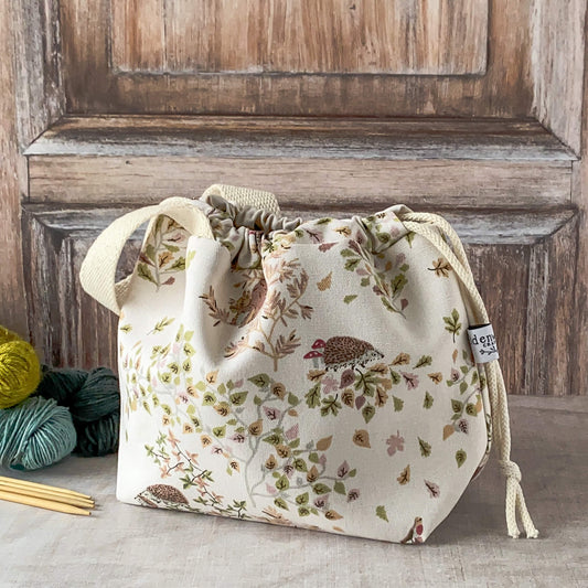 A small knitting project bag with a soft woodland design, featuring hedgehogs, birds, and delicate leaves in pastel hues. The bag has a drawstring closure and sturdy fabric handles. It is placed on a neutral surface with nearby yarn and knitting needles.