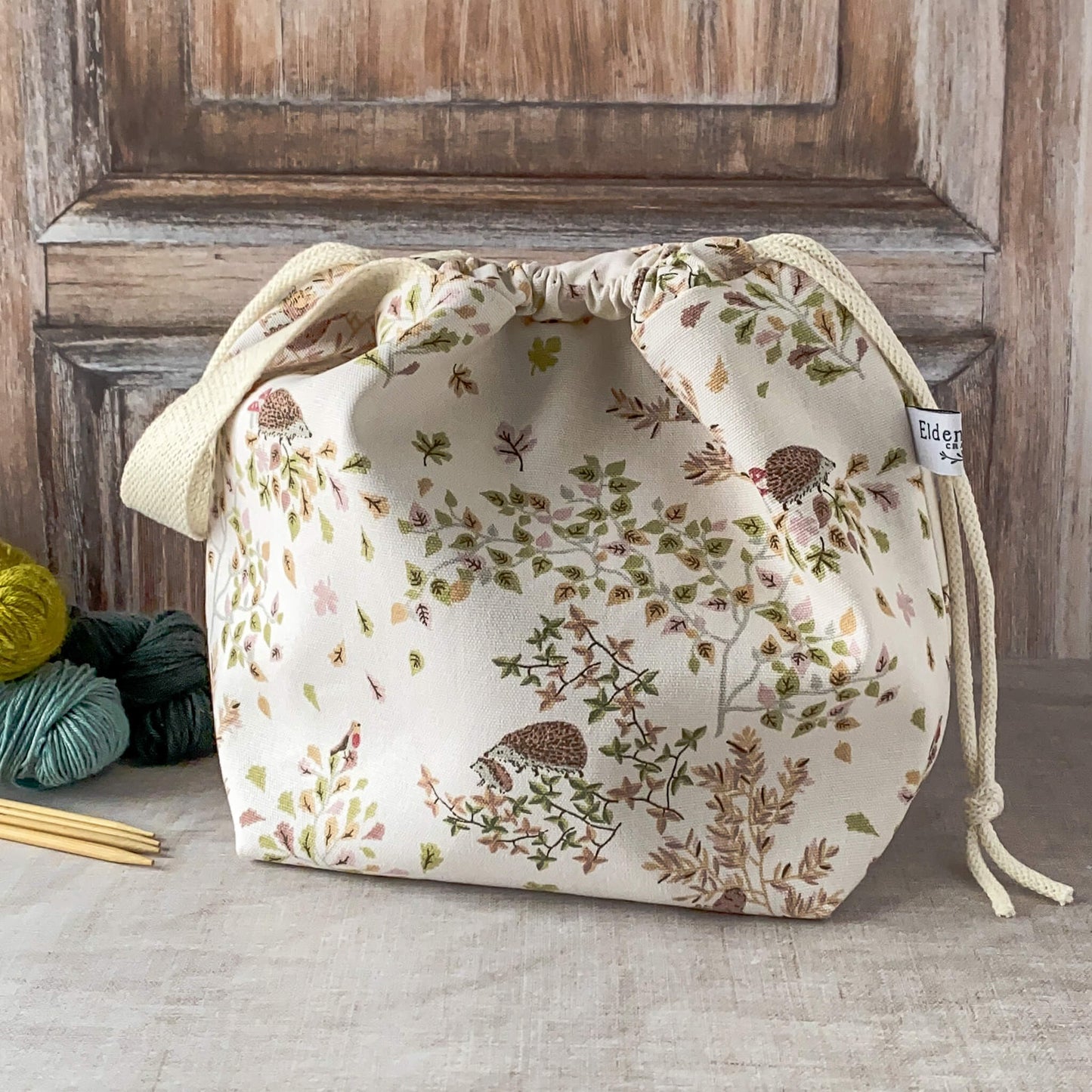 A medium-sized knitting project bag with a charming woodland design, featuring hedgehogs, birds, and leaves in pastel hues. The bag has a drawstring closure with fabric handles, and nearby are knitting needles and colorful skeins of yarn on a neutral surface.