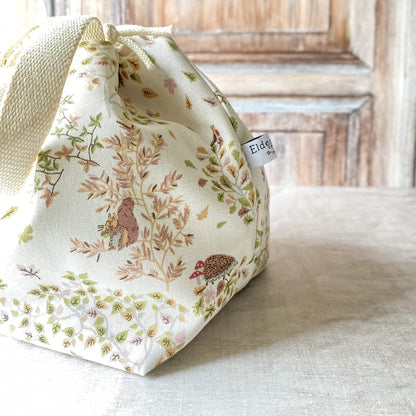 A close-up of a medium-sized knitting project bag adorned with soft illustrations of woodland creatures, including birds and hedgehogs, set among delicate foliage. The bag features fabric handles and a drawstring closure, with knitting needles and yarn visible next to it.