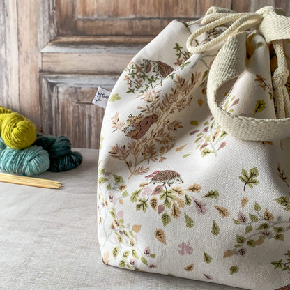 A beautifully designed medium-sized knitting project bag with woodland motifs like hedgehogs and birds surrounded by gentle foliage. The bag is cinched with a drawstring and has sturdy handles. Skeins of yarn and knitting needles are placed on the wooden surface beside it.