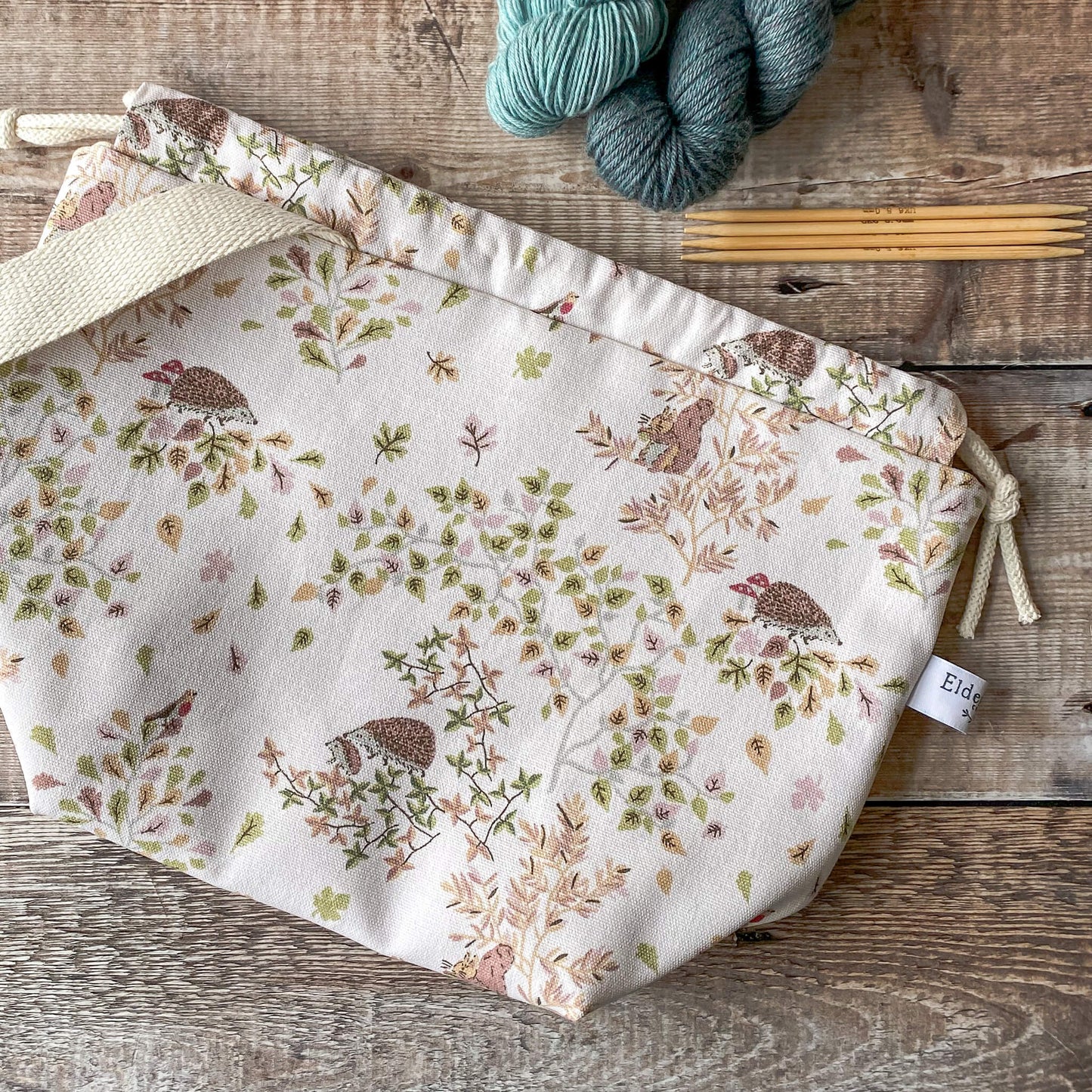 A medium-sized knitting project bag featuring a woodland friends print with hedgehogs, birds, and leaves in soft, earthy tones. The bag is closed with a drawstring and has fabric handles. Nearby, knitting needles and yarn in various colors are visible on the wooden surface.