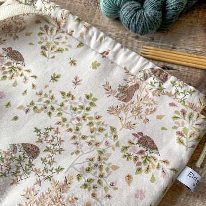 A detailed view of a medium-sized knitting project bag with a whimsical woodland scene, showcasing hedgehogs, birds, and autumn leaves. The bag features a drawstring closure and sturdy handles. Nearby, knitting needles and colorful skeins of yarn are set on a wooden background.