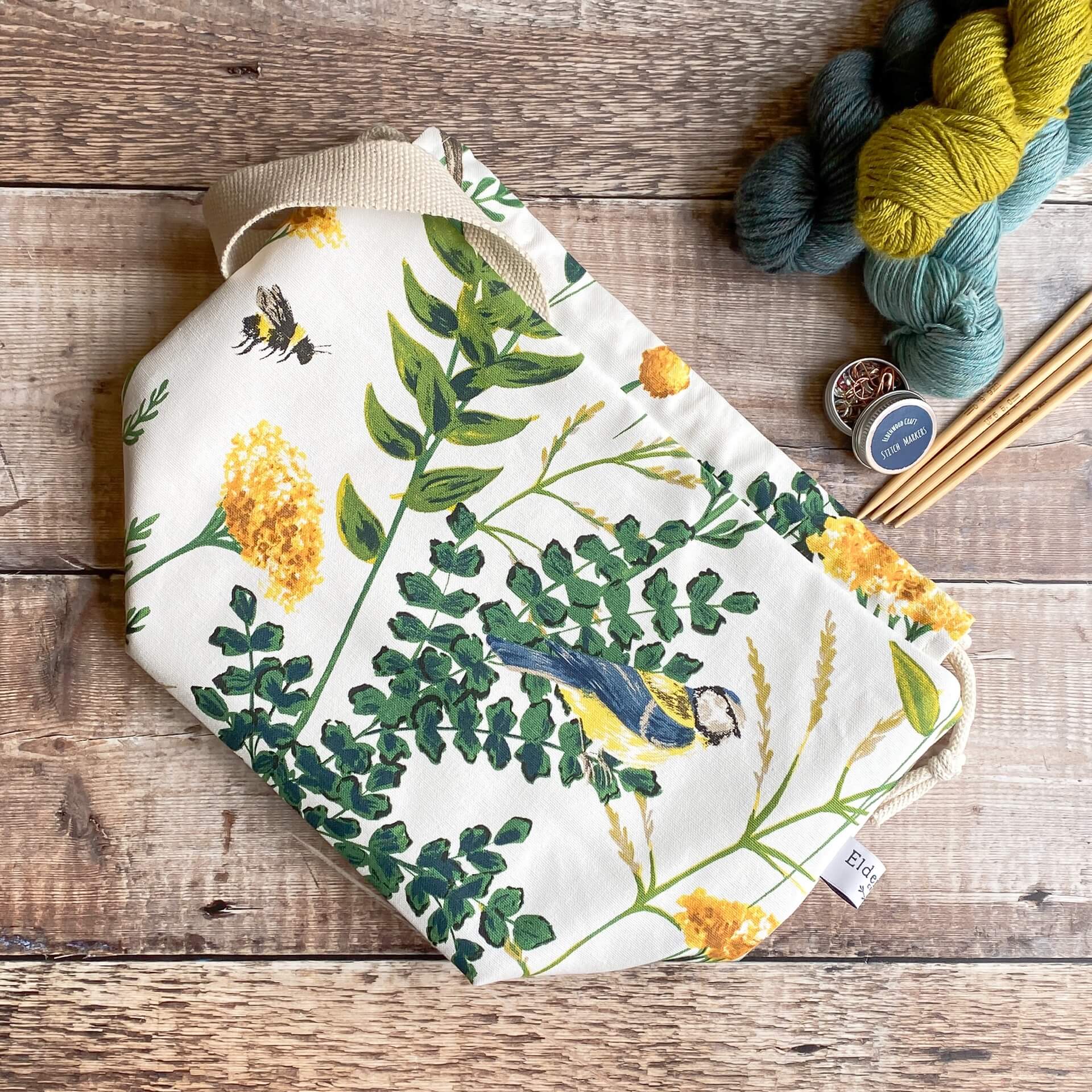 A drawstring bag lies flat on a wooden table next to some hanked up yarn, stitch markers and wooden knitting needles. The bag has a print of a summery scene featuring blue tit birds and bumble bees. 