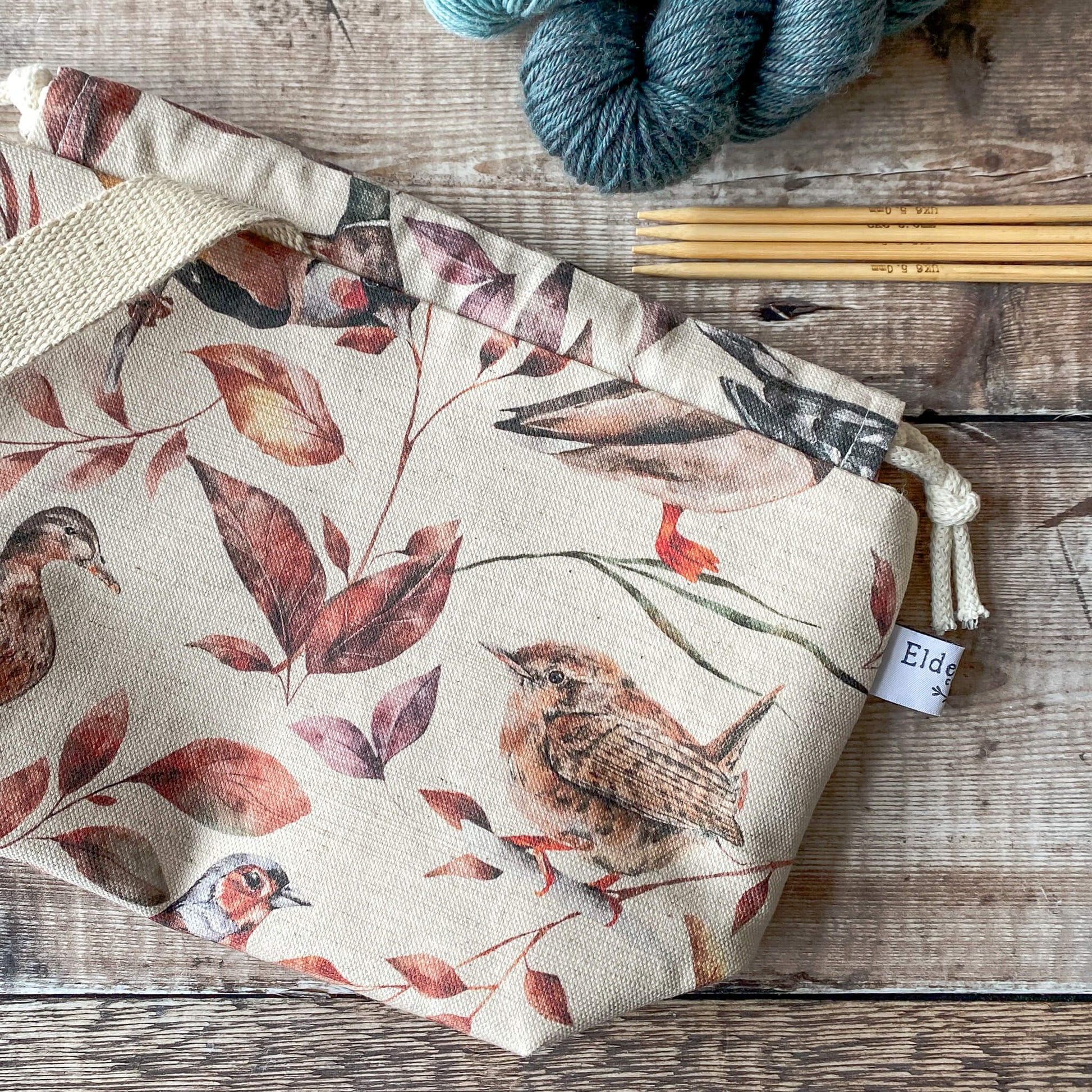 A close-up of a small knitting project bag with a soft woodland design featuring birds and colorful autumn leaves. The bag is closed with a drawstring and has sturdy fabric handles. It sits on a wooden surface, surrounded by skeins of yarn and knitting needles.