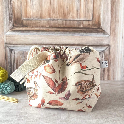 A medium-close-up of a small bag showcasing woodland bird and leaf prints in warm autumn hues. The bag is cinched with a drawstring and has a sturdy fabric handle. It is placed on a neutral surface with yarn and knitting needles nearby.