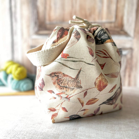 A small knitting project bag with a woodland bird design, featuring soft tones of autumn leaves and a bird perched on a branch. The bag has a drawstring closure with fabric handles. Nearby are skeins of yarn and knitting needles on a neutral background.