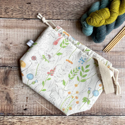 A knitting project bag lies flat on a wooden table next to three skeins of yarn and some wooden knitting needles. The bag is made from a print covered in wildflowers. 