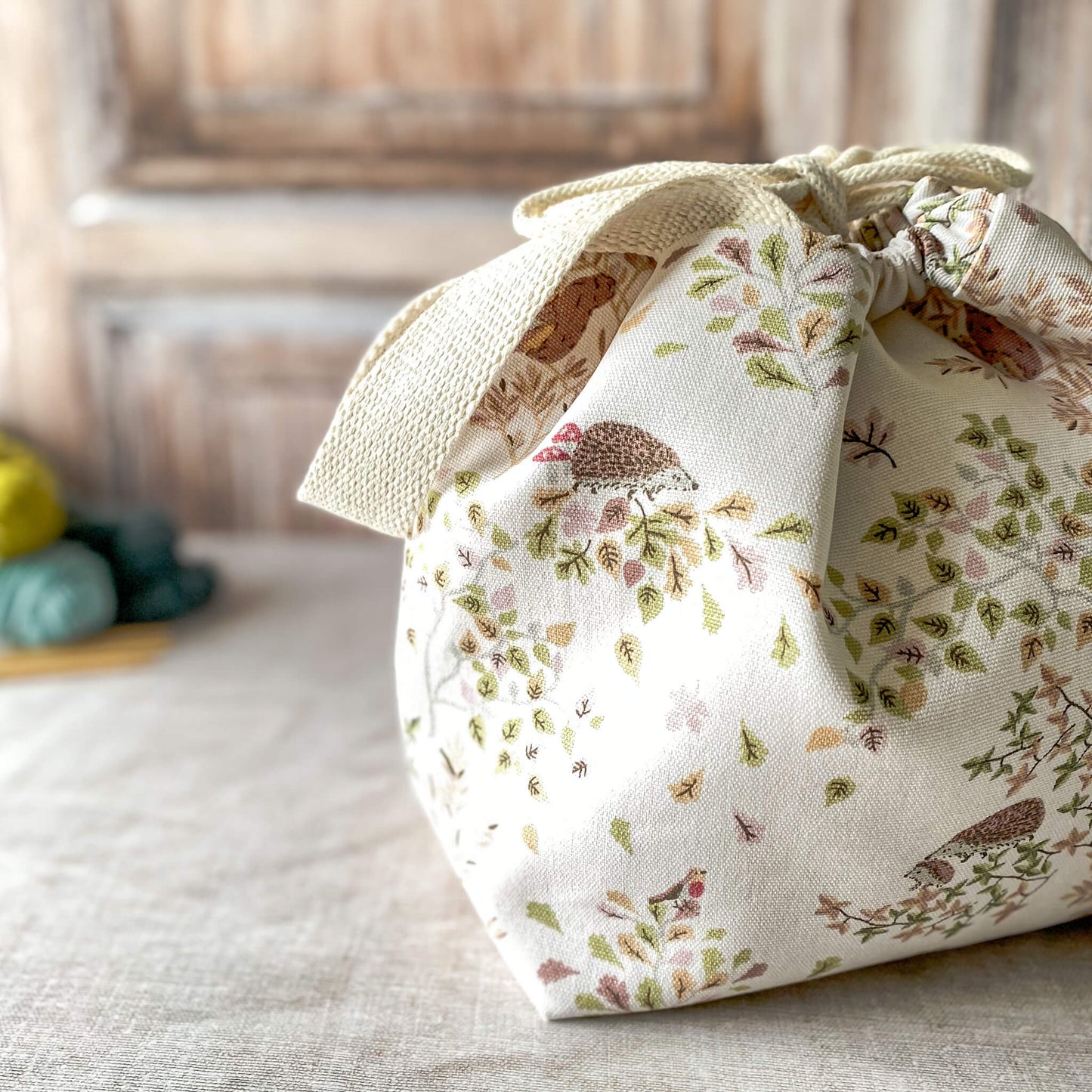 A close up of a small knitting project bag with a soft woodland design, featuring hedgehogs, birds, and delicate leaves in pastel hues. The bag has a drawstring closure and sturdy fabric handles. It is placed on a neutral surface with nearby yarn and knitting needles.