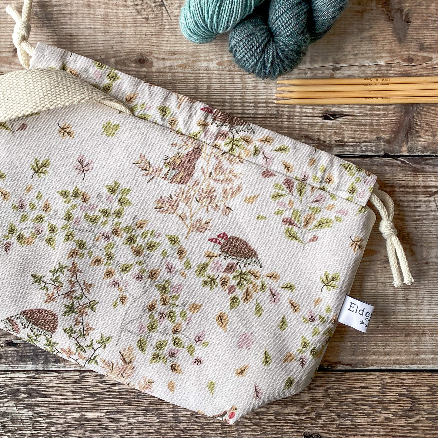 A close-up of a small knitting project bag adorned with woodland creatures such as hedgehogs and birds, surrounded by soft foliage. The bag features a drawstring closure and fabric handles. Knitting needles and yarn are visible on the wooden surface next to the bag.