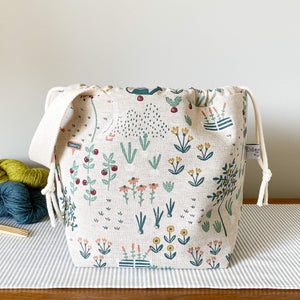 A drawstring bag sits on a table top. Behind the bag are some skeins of yarn in different colours neatly arranged. Also in view are some wooden knitting needles. The bag is at the centre of the image and is made from a fabric that depicts a cottage garden. 