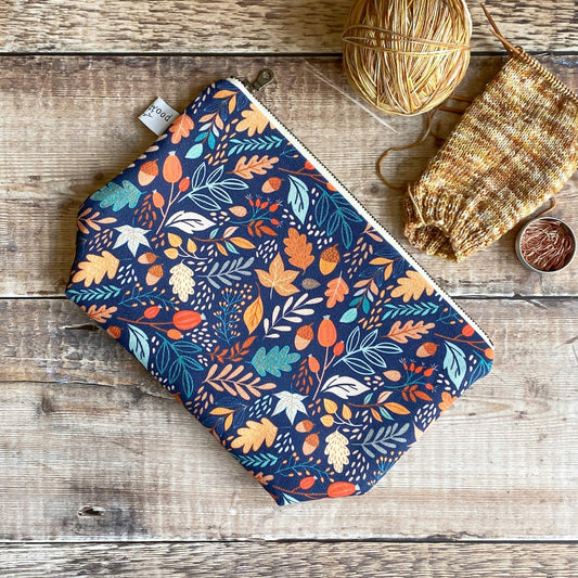 A small zipped pouch which features a print of lots of small leaves and acorns sits on a wooden table next to some in progress knitting and a tin of stitch markers. 