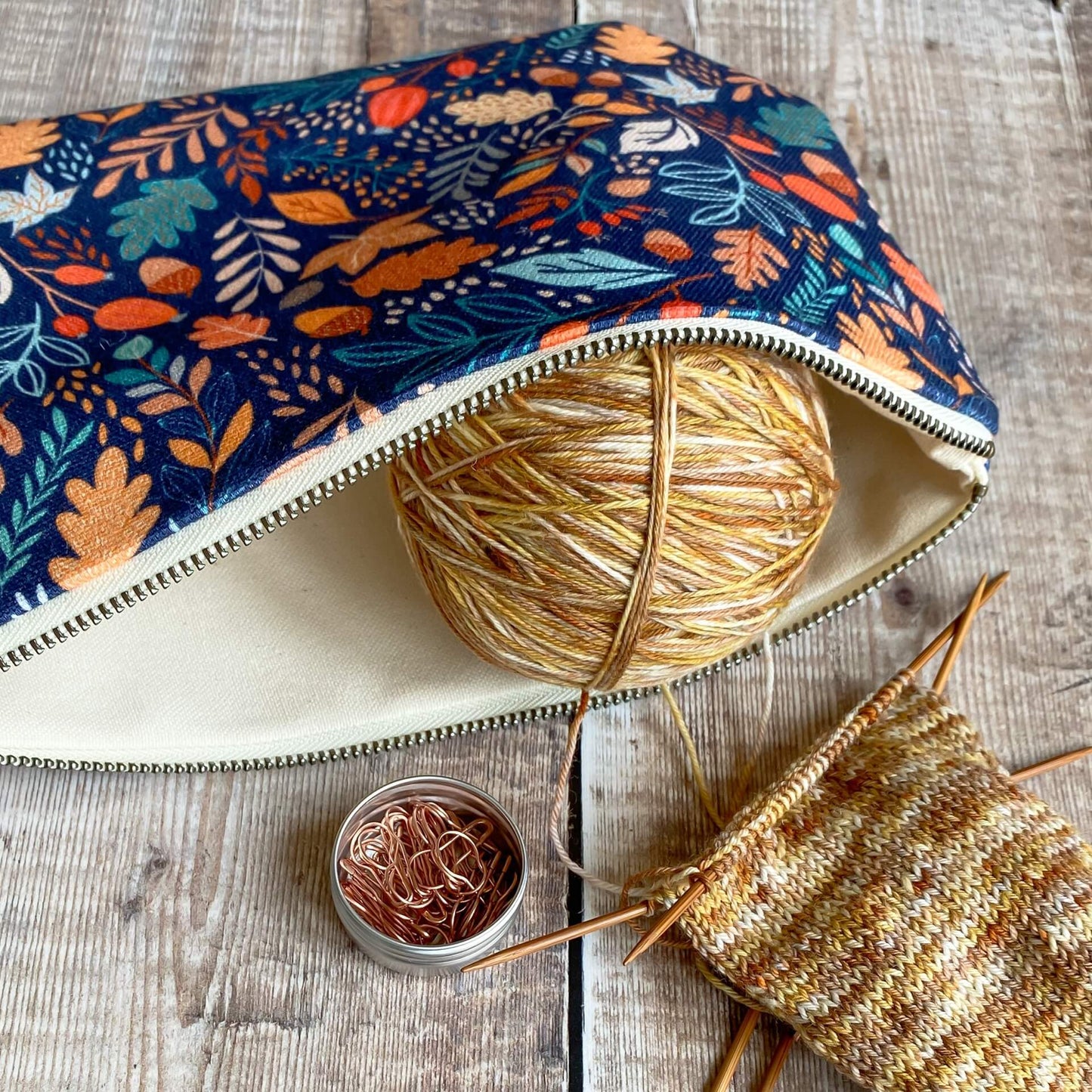 A zipped pouch lies open. In the opening a ball of yarn in brown tones sits. The yarn is attached to an in progress knitting project. Also next to the pouch is a tin of stitch markers. 