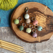Load image into Gallery viewer, Six stitch markers and a progress keeper for knitting sit on a wooden block of wood. The wood is placed on a wooden table top which has a blue cloth laid over it. Next to the stitch markers is a skein of green yarn and four wooden knitting needles. The stitch markers are made from wool felt balls and are in faded summer colours. 