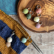Load image into Gallery viewer, Five stitch markers for knitting sit on a wooden block of wood. The wood is placed on a wooden table top which has a blue cloth laid over it. Next to the stitch markers is a knitting project in progress. Attached to the knitting project is one stitch marker and a progress keeper. The stitch markers are made from wool felt balls and are in faded summer colours. 