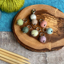 Load image into Gallery viewer, Six stitch markers and a progress keeper for knitting sit on a wooden block of wood. The wood is placed on a wooden table top which has a blue cloth laid over it. Next to the stitch markers is a skein of green yarn and four wooden knitting needles. 