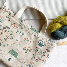 Load image into Gallery viewer, A large drawstring project bag for knitting lies flat on a linen covered table. Next to the bag are two skeins of yarn. One is blue and the other green. Next to the yarn lies four wooden knitting needles. 