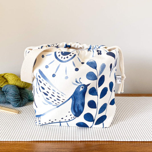 A knitting project bag adorned with a vibrant indigo folk art print rests on a wooden table. Behind the bag, three colorful skeins of yarn stand in an orderly fashion.