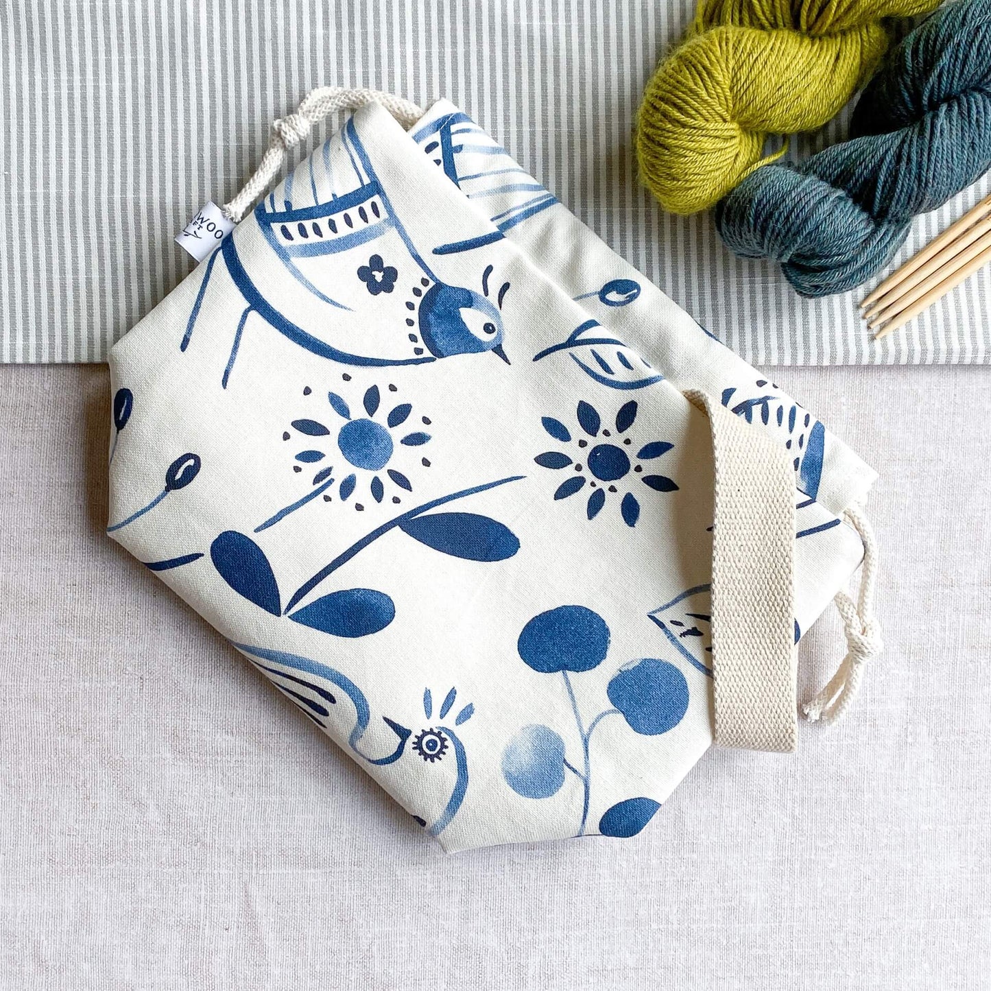 An indigo-colored folk art print knitting project bag is lying flat on a table top. The bag is rectangular in shape and made of fabric. It features a vibrant folk art image on its surface. Just in view are two skeins of yarn and four wooden knitting needles. 