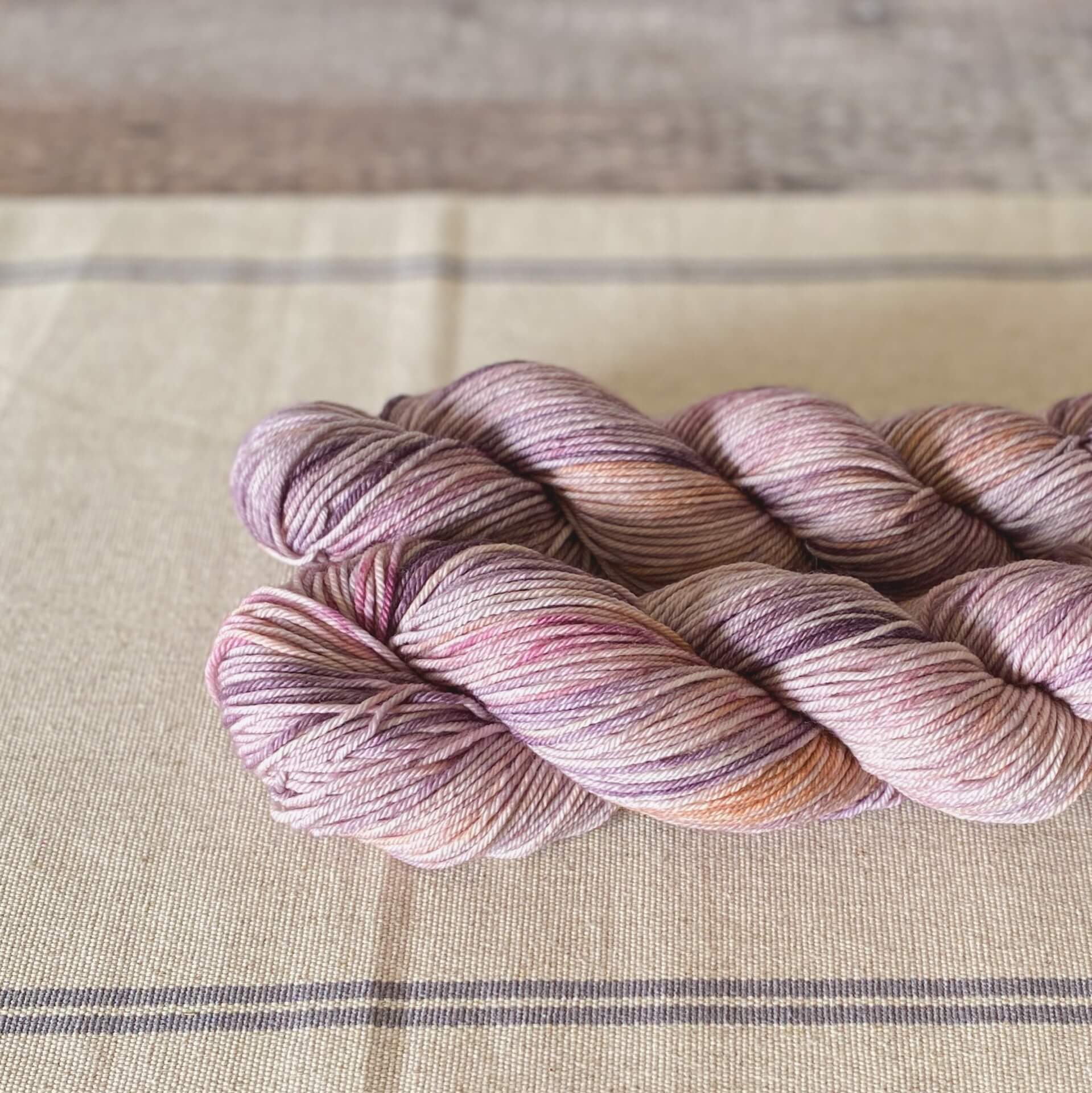 Two skeins of yarn lie on a linen table runner. The yarns have coloured in pinks and purples. 