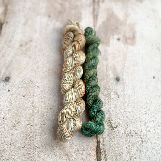 Two skeins of yarn sit on a wooden table. One is slightly bigger than the other. The larger consists of colours such as terracotta and beige with sprinkels of blue. The smaller skein is a green colour. Together they are reminiscent of beaches on the south coast of Devon. 