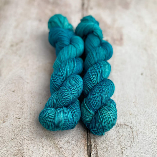 Two skeins of hand dyed yarn sit together on a wooden table. The consist of shades of tealy blue and are reminiscent of the colurs of an exotic lagoon. 