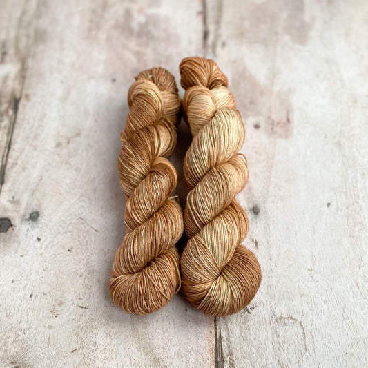 Two skeins of hand dyed yarn sit next to each other on a wooden table. They are a milky brown colour and remind one of frothy coffee, hence the name of the colourway. 