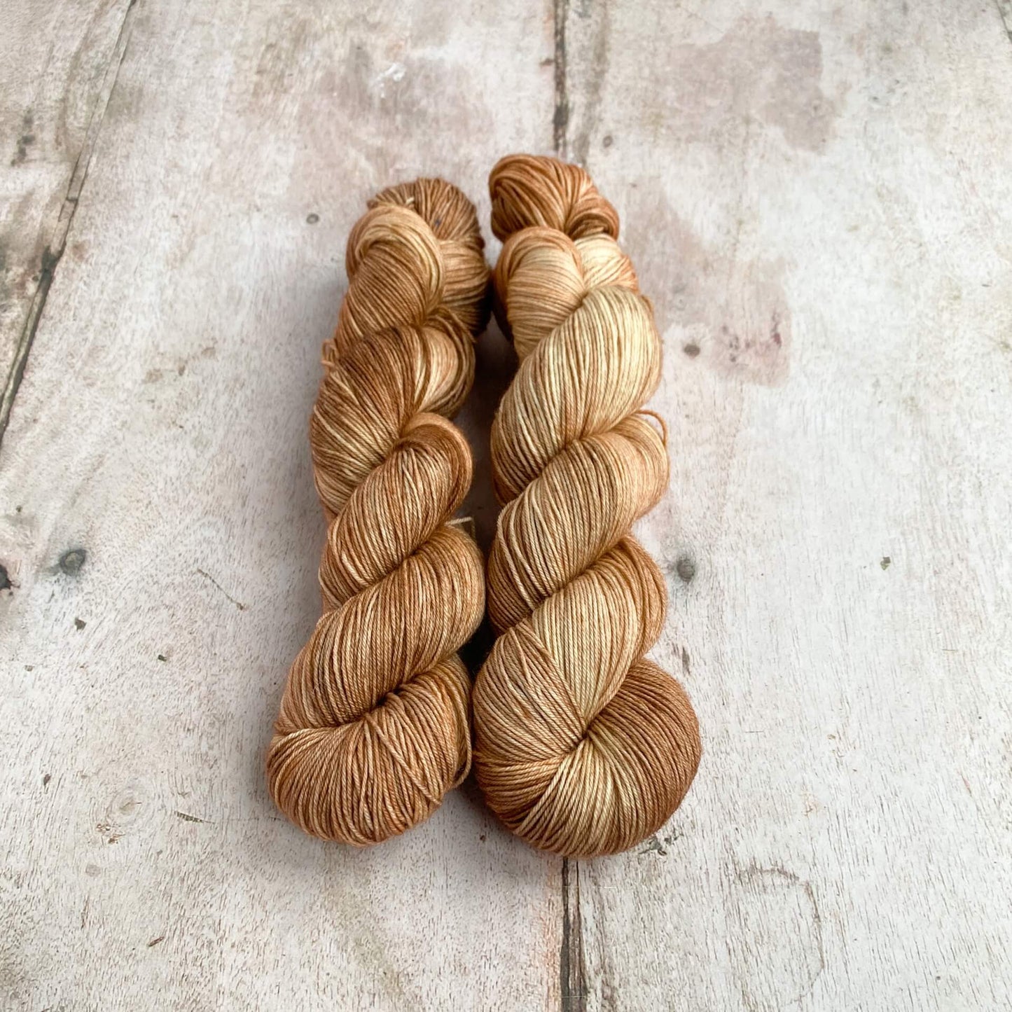 Two milky brown hand dyed skeins of yarn sit next to each other on a wooden table. They are wound into hanks. 