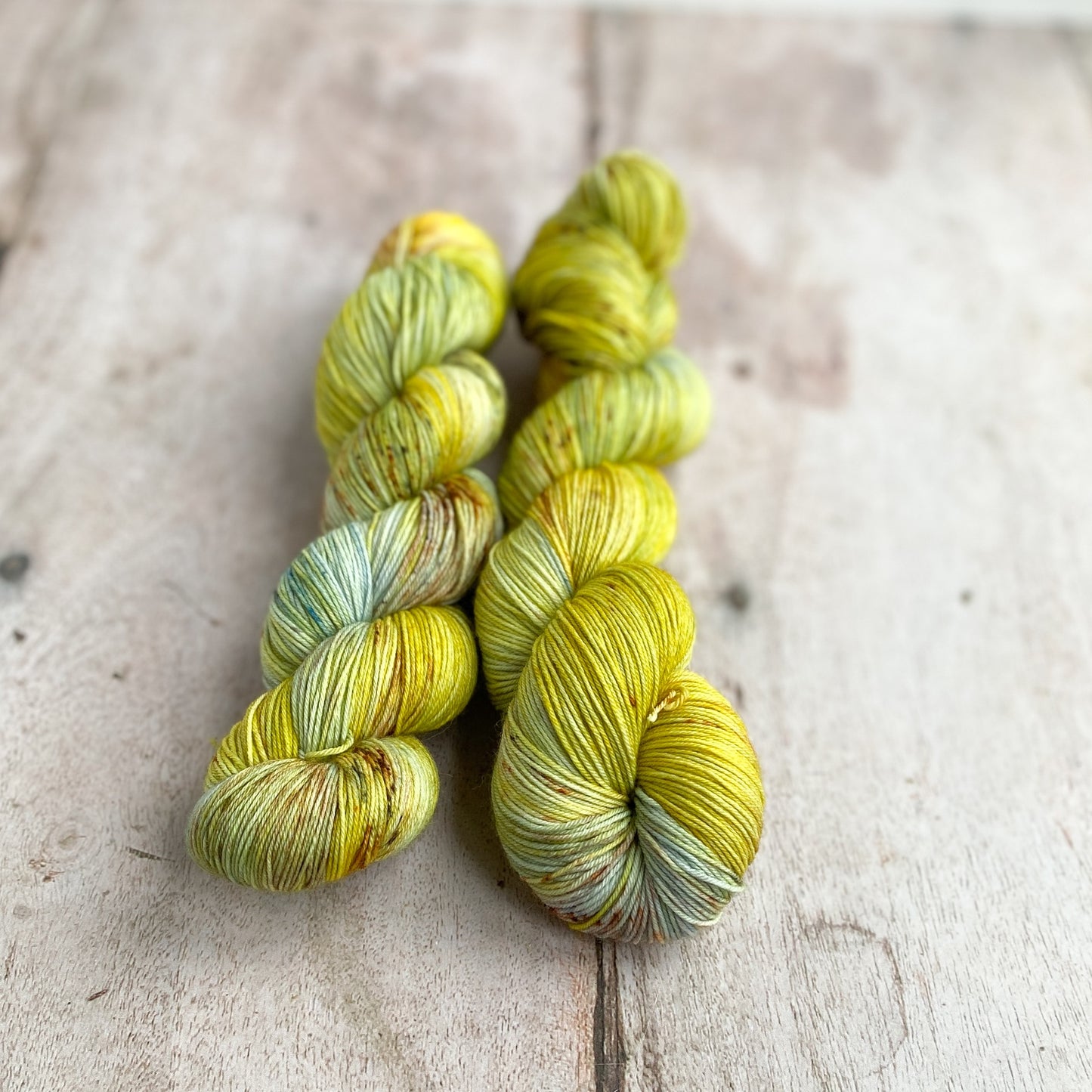 Two skeins of hand dyed yarn sit next to each other on a wooden table. They are coloured with shades of golden green, pale blue and lots of chestnut sprinkles. 