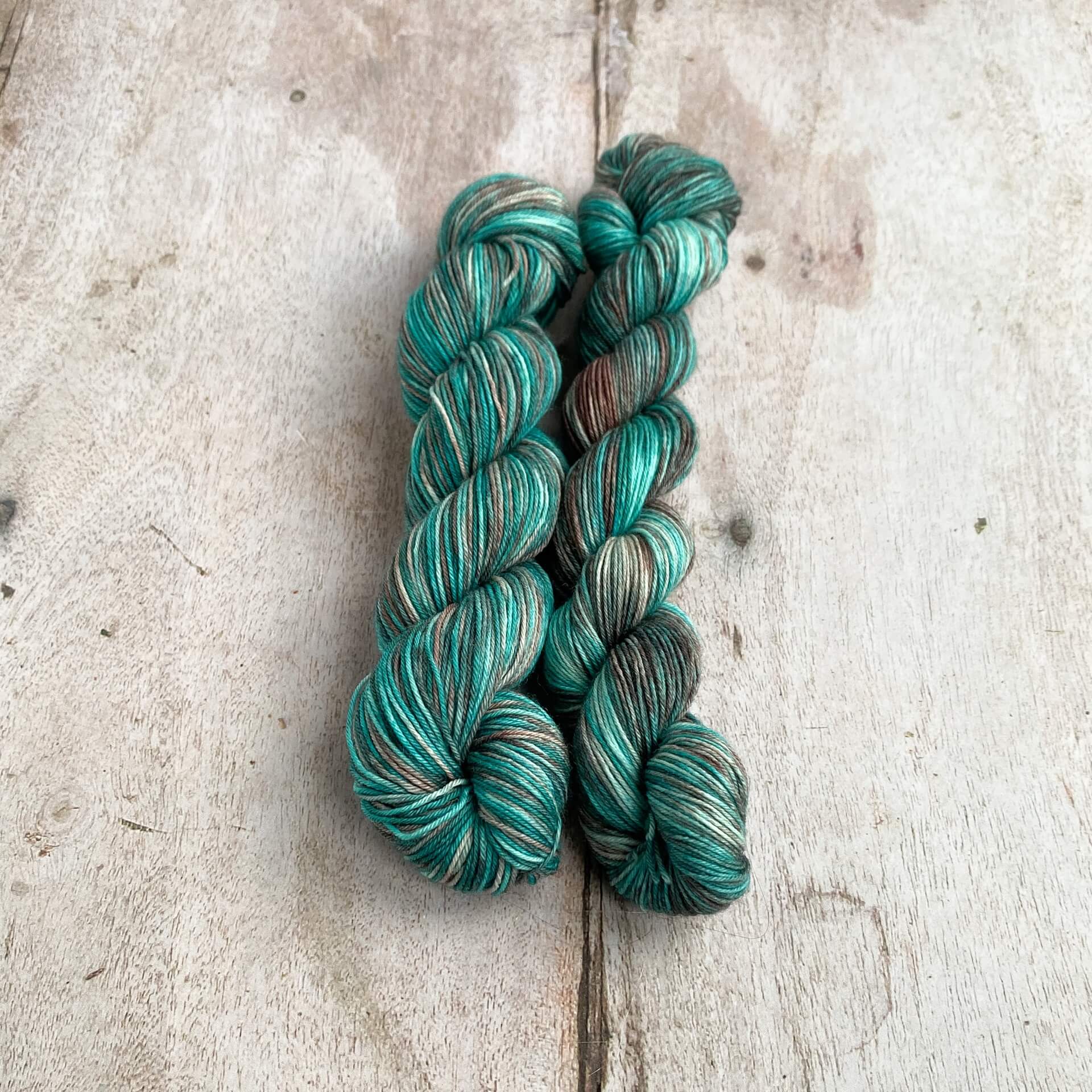 Two skeins of yarns dyed with teals and browns sit togther on a wooden table. 