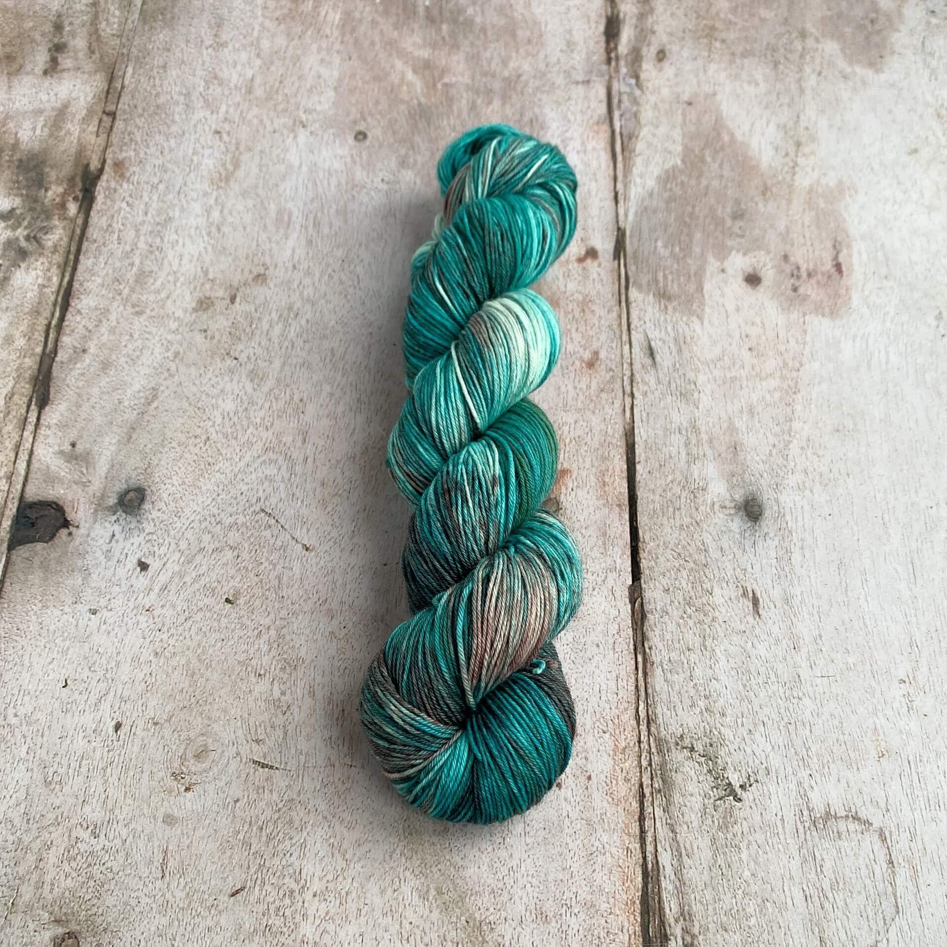 A skein of yarn lies on a wooden table. It is coloured with teals and browns. 