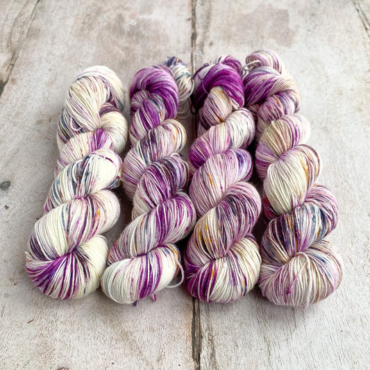 Four skeins of hand dyed yarn sit on a wooden table. They are dyed using purple dyes along with golds and blues. 