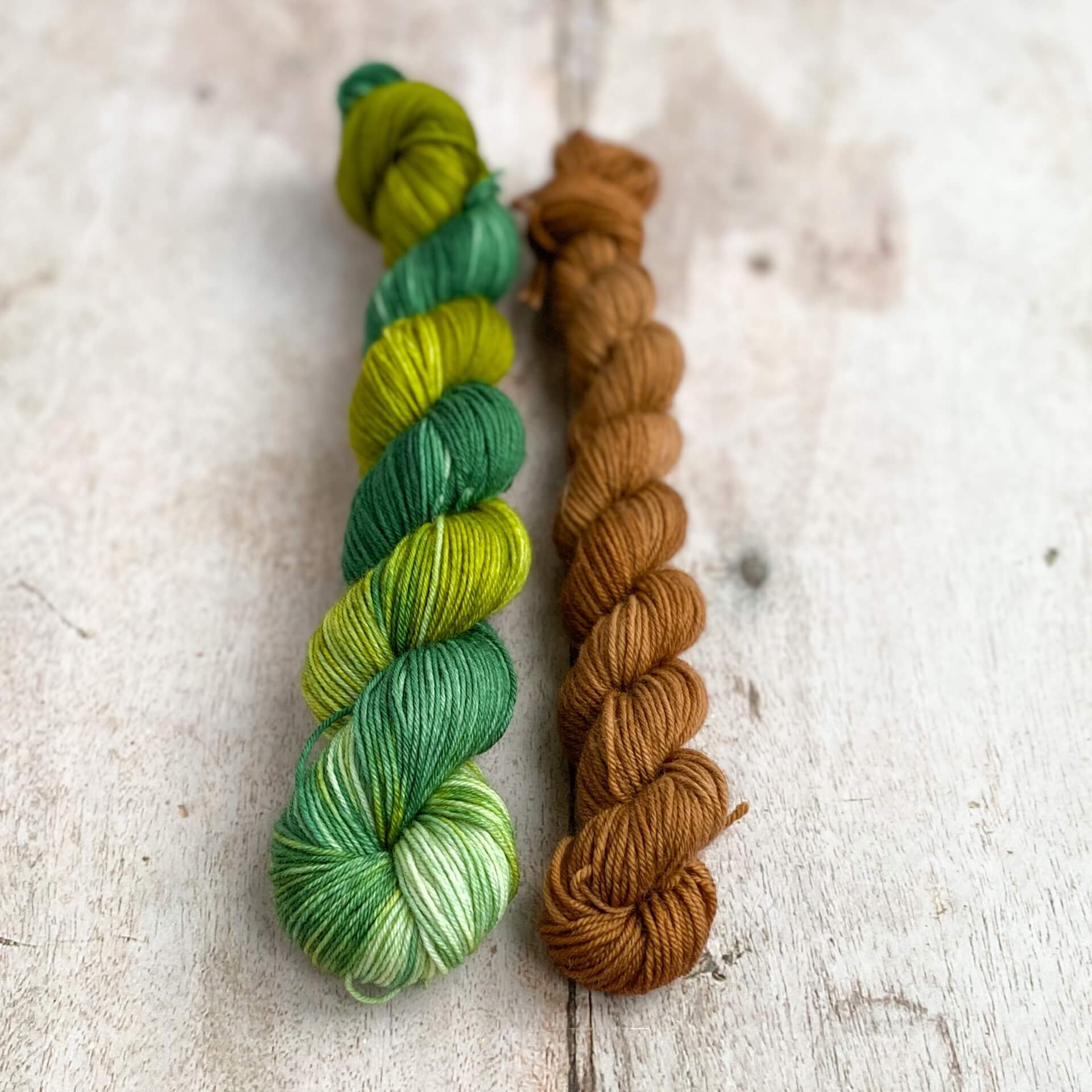 Two skeins of yarn on a wooden table. One is dyed with green dyes and the other is dyed with a brown dye. Together they look like a fir tree in winter. 