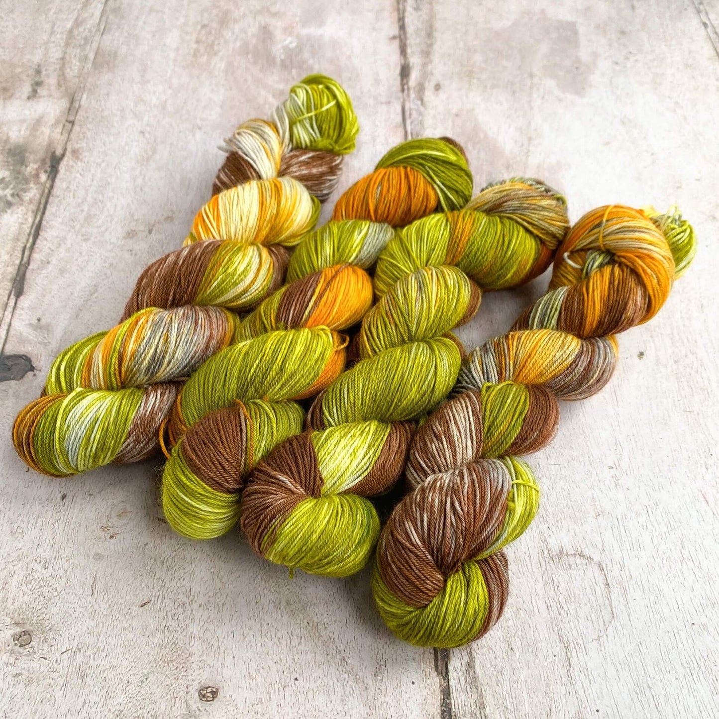 Four skeins of hand dyed yarn lie on a wooden table. The colours in the skeins are browns, greens and oranges and are reminiscent of the colours of autumn.