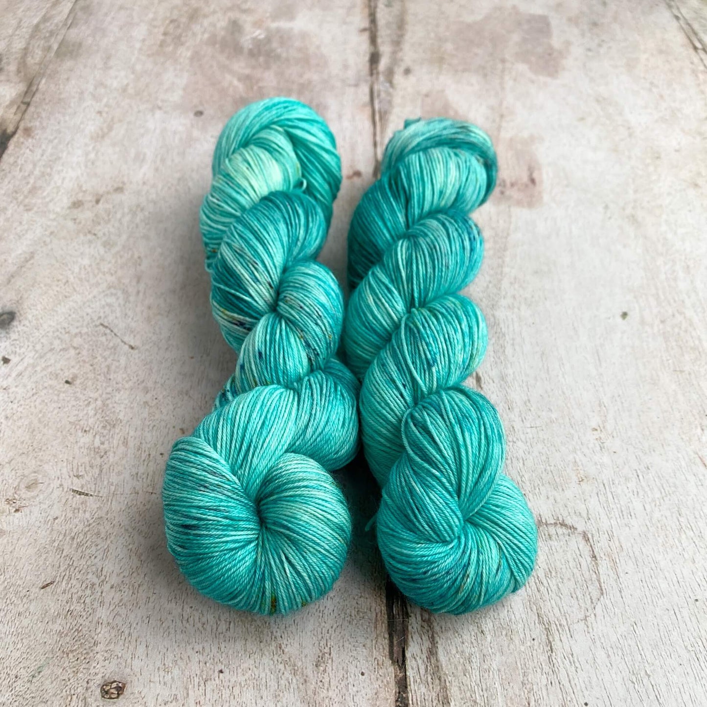 Two skeins of hand dyed yarn sit on a wooden table. They are dyed with an aqua blue dye and have slight sprinkles added for interest. 