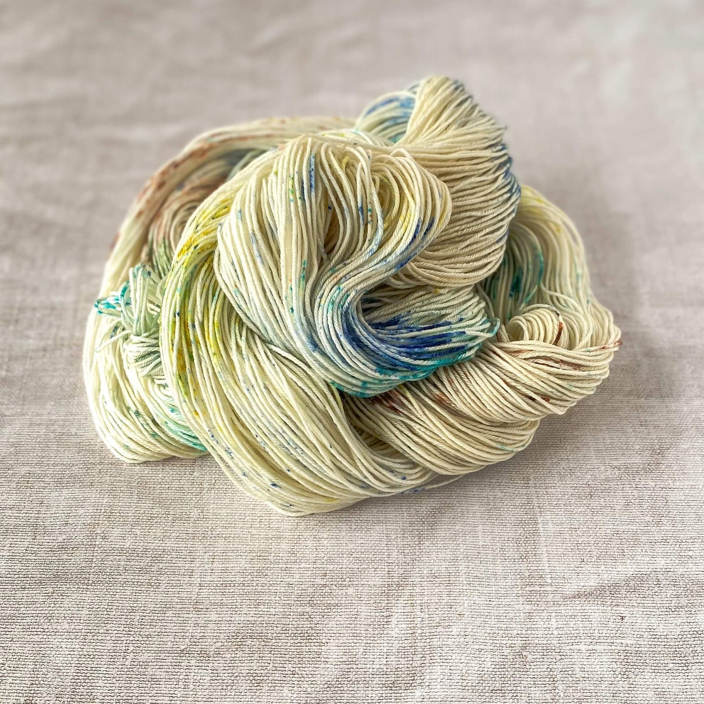 A loosely coiled hank of hand-dyed yarn in a soft cream color with speckles of blue, green, yellow, and brown, resting on a neutral fabric background.