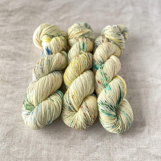 Three skeins of hand-dyed yarn in a soft cream color with speckles of blue, green, yellow, and brown, neatly twisted and arranged on a neutral fabric background.