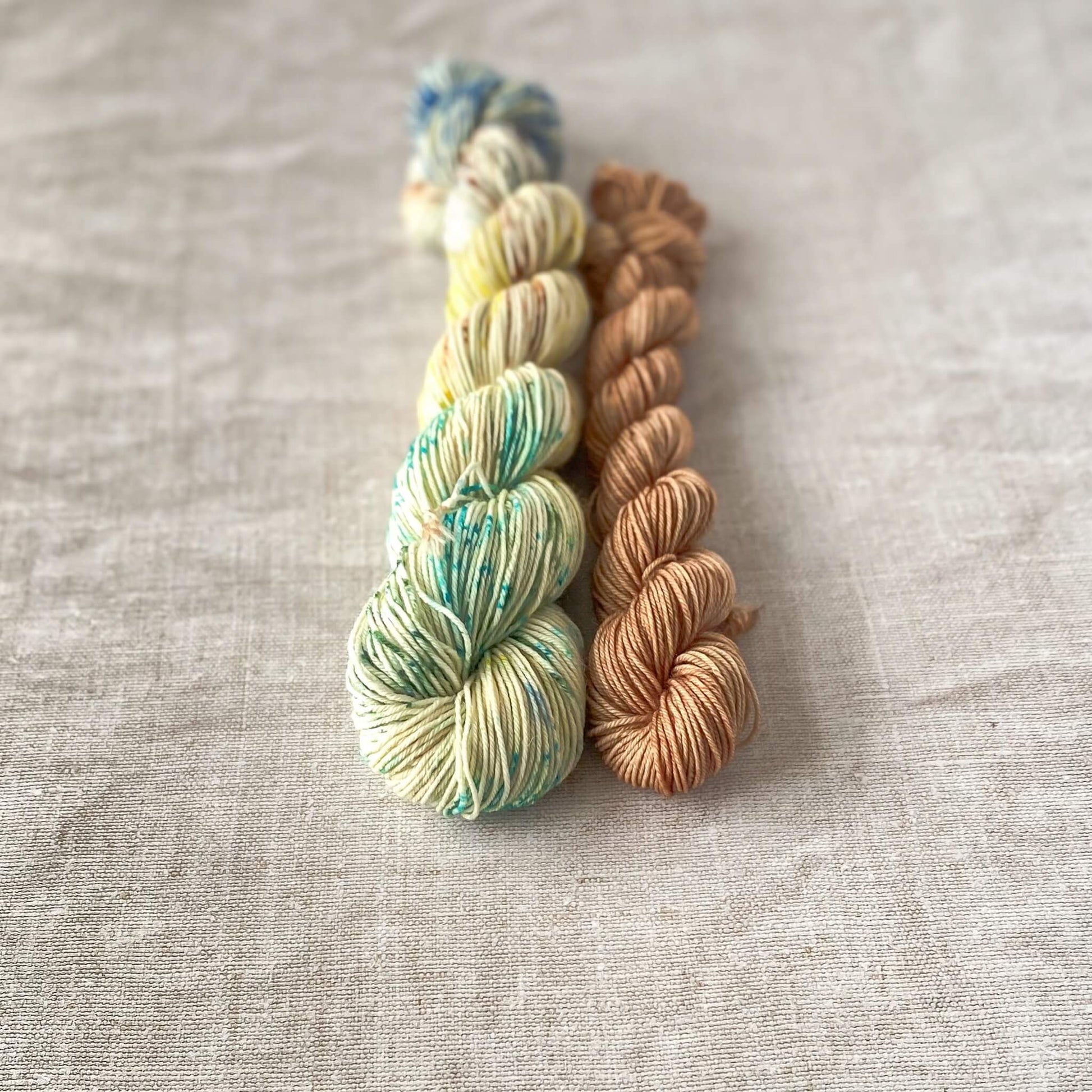 Two skeins of hand-dyed yarn lying parallel on a neutral fabric background. The larger skein is cream-colored with speckles of blue, green, yellow, and brown, while the smaller skein is a warm, earthy brown.